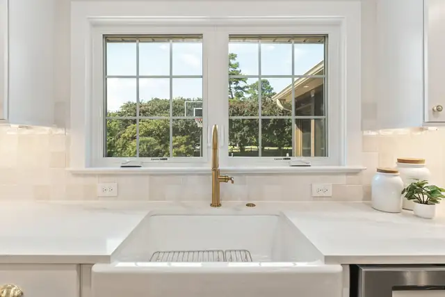 White farmhouse sink with a brass faucet under a double window, with white cabinets and a tiled backsplash