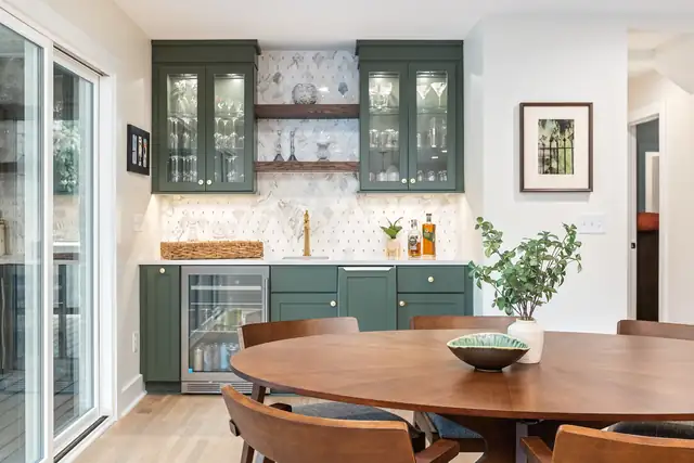 Dining area beside a green built-in bar with glass-front cabinets, a beverage fridge, wood shelves and a marble mosaic backsplash