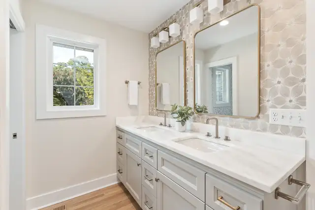 Bathroom with a long gray double vanity, a white stone counter, brass faucets and a patterned tile wall behind two mirrors