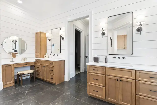 Bathroom with white shiplap walls, wood vanities with a seated makeup station, black framed mirrors and dark stone-look floor tile